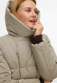 Manteau matelassé beige avec un col montant et une grande capuche. Comprend des poignets en maille côtelée marron et une texture matelassée. Finition extérieure lisse et douce.