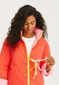 Prošívaná oranžová bunda s růžovým límcem, žlutým zipem a barevnými stahovacími šňůrkami. Má diamantový vzor a hladkou strukturu.