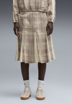 Personne portant une jupe plissée à carreaux beige, des chaussettes blanches Puma et des baskets beiges avec semelles en gomme, debout devant un fond gris.