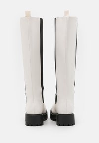 Botas brancas até ao joelho com painéis laterais elásticos pretos e solas grossas pretas apresentam um acabamento em couro suave e uma alça na parte superior.