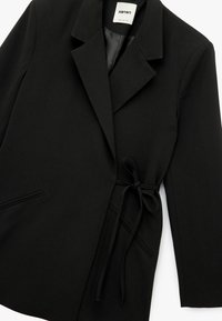 Blazer noir en tissu lisse, avec col cranté, fermeture à nouer à l'avant et deux poches latérales avec des détails de piqûre subtils.