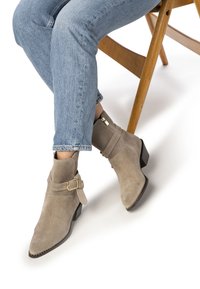 Bottines en suède tan avec une fermeture éclair sur le côté, une sangle contrastante et un détail de boucle dorée, et un talon bas et épais. Portées avec un jean bleu clair.