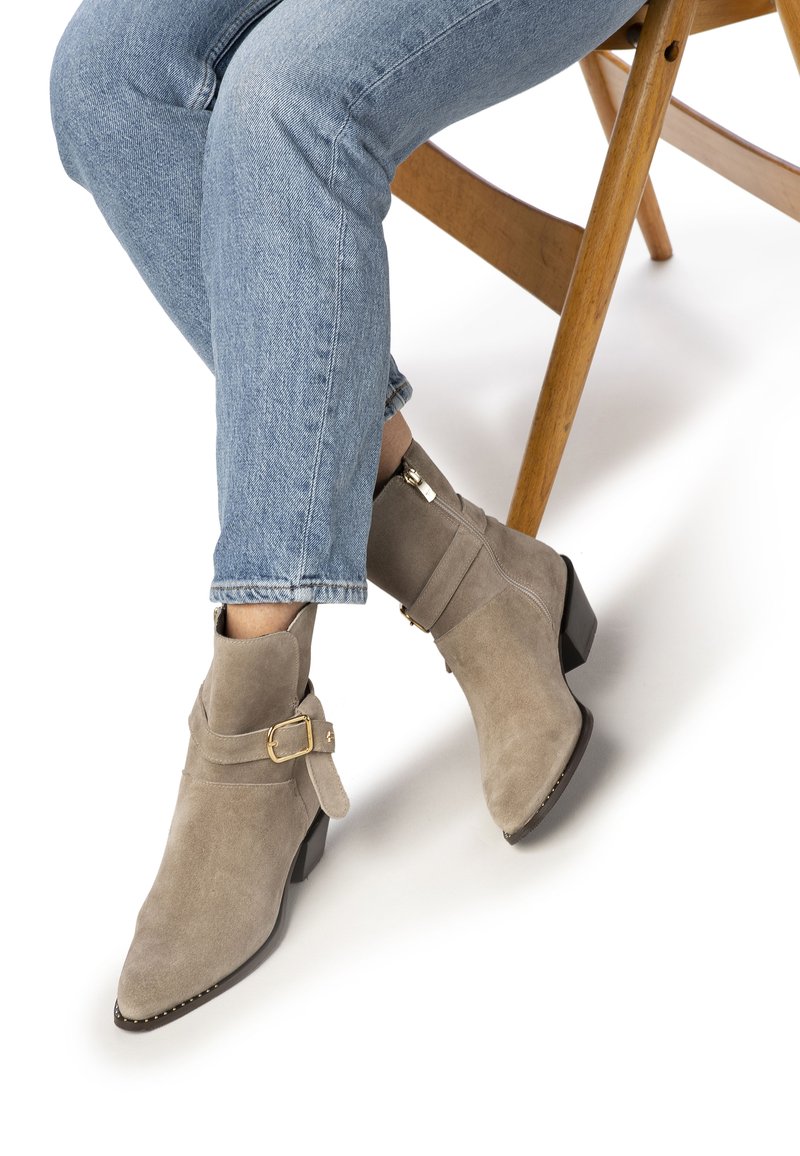 Bottines en suède tan avec une fermeture éclair sur le côté, une sangle contrastante et un détail de boucle dorée, et un talon bas et épais. Portées avec un jean bleu clair.