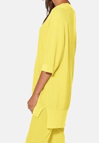 Žlutá pletená tunika s volnějším střihem, krátkými rukávy a bočními rozparky pro snadnější pohyb. Texturovaná látka a jednobarevný design.