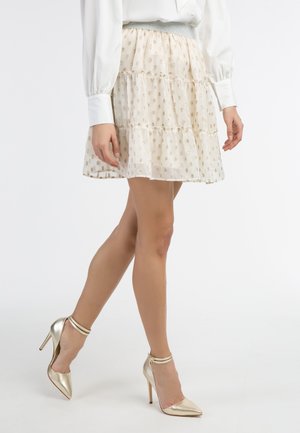 Frau trägt einen beigen Blumenrock, weiße Bluse und goldene High-Heel-Riemchenschuhe und steht mit einem Bein über das andere gekreuzt.