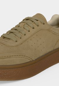 Gros plan d'une basket en daim beige avec des lacets assortis et une semelle en caoutchouc marron texturée sur fond blanc.