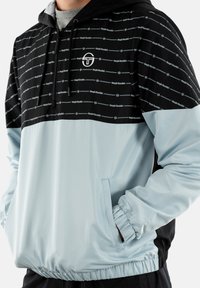 Uomo che indossa una giacca con cappuccio Sergio Tacchini nera e azzurra chiara, con zip e mano in tasca, che mostra i dettagli del busto e delle maniche.