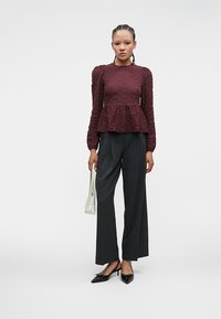 Top peplum testurizzato color bordeaux con maniche lunghe, abbinato a pantaloni neri a vita alta e décolleté nere a punta. Gli accessori includono una piccola borsa bianca.