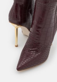 Burgundin väriset korkokengät nilkkasaappaana, joissa on kiiltävä krokotiilikuvio, terävä kärki ja metallinhohtoinen kultainen stilettokorko, jossa on pieni kumipää.