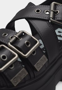 Μαύρο δερμάτινο σανδάλι με δύο ρυθμιζόμενες ασημένιες αγκράφες και παχιά σόλα, με μεταλλικές ετικέτες με το λογότυπο στα λουριά.