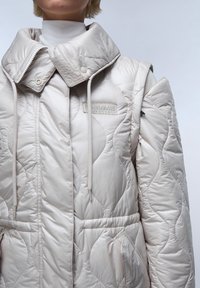 Prošívaná béžová syntetická bunda s vysokým límcem, stahovacími šňůrkami a předními kapsami. Má texturovaný, polstrovaný design a detail s logem.