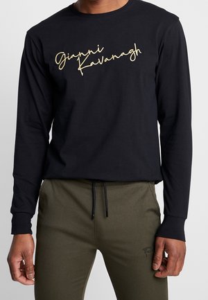 Chemise noire en coton à manches longues présentant du texte doré en cursive "Gianni Kavanagh" sur le devant. Texture douce avec une coupe décontractée, associée à un pantalon olive.