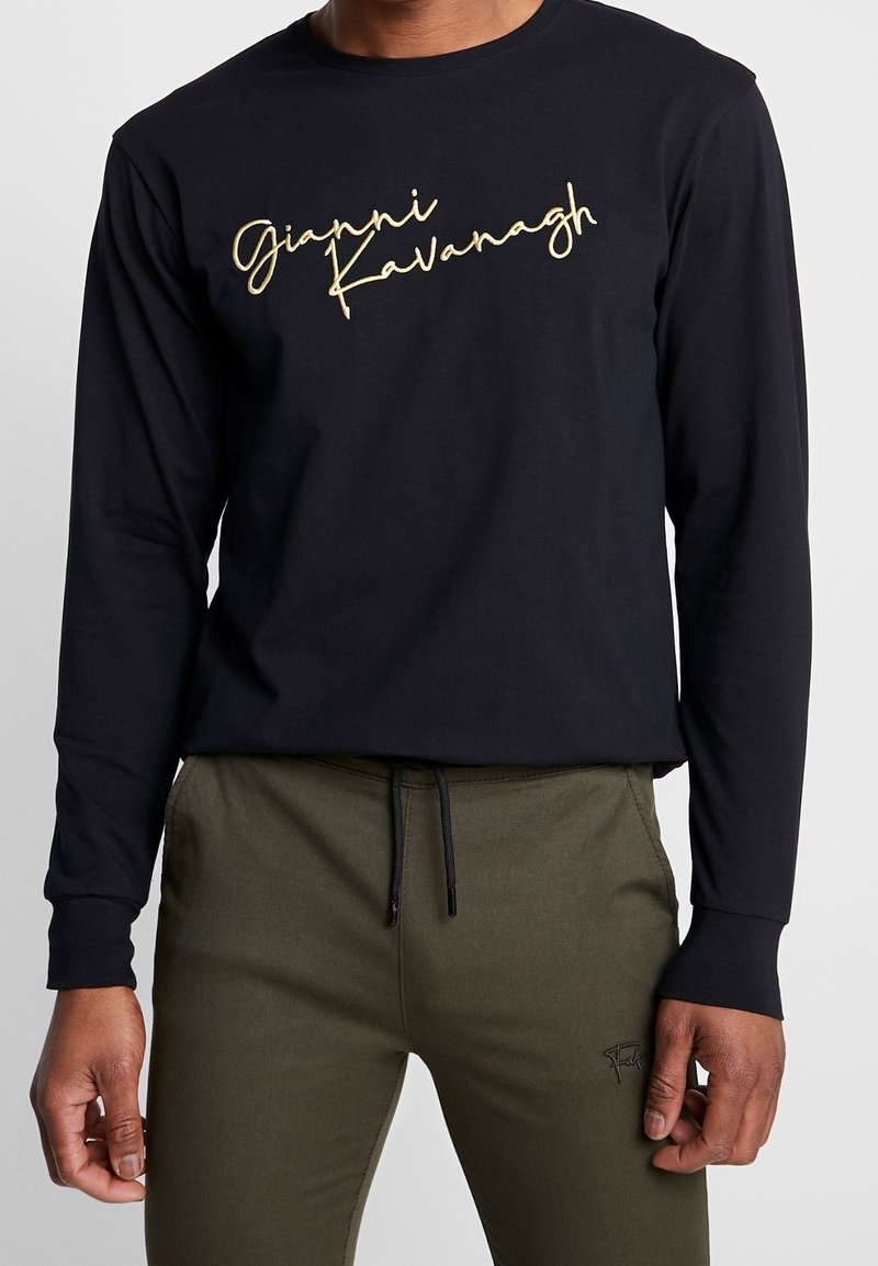 Černá košile s dlouhým rukávem vyrobená z bavlny, na přední straně s nápisem "Gianni Kavanagh" v zlatém kurzívě. Měkká textura a volný střih, doplněná olivovými kalhotami.