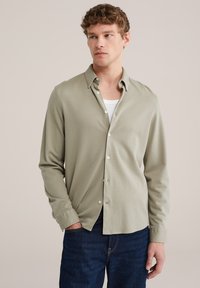 Hellgrünes Hemd mit Knopfleiste, strukturierter Stoff, figurbetonter Schnitt und langen Ärmeln, kombiniert mit dunkelblauen Jeans. Sichtbare Kragenknopf-Details.