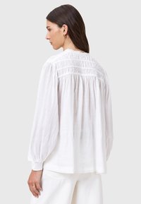 Blusa bianca con maniche lunghe a sbuffo, dettagli raccolti a trama nella parte posteriore e una vestibilità ampia e fluida realizzata in tessuto leggero.