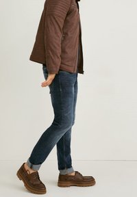 Mann in brauner Jacke, dunkelblauen hochgekrempelten Jeans und braunen Schnürschuhen, seitlich stehend vor einem schlichten hellen Hintergrund.