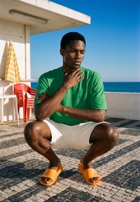 Jeune homme en chemise verte et short blanc accroupi sur une terrasse carrelée au bord de la mer, portant des sandales orange, avec des chaises rouges et un parasol rayé à proximité.