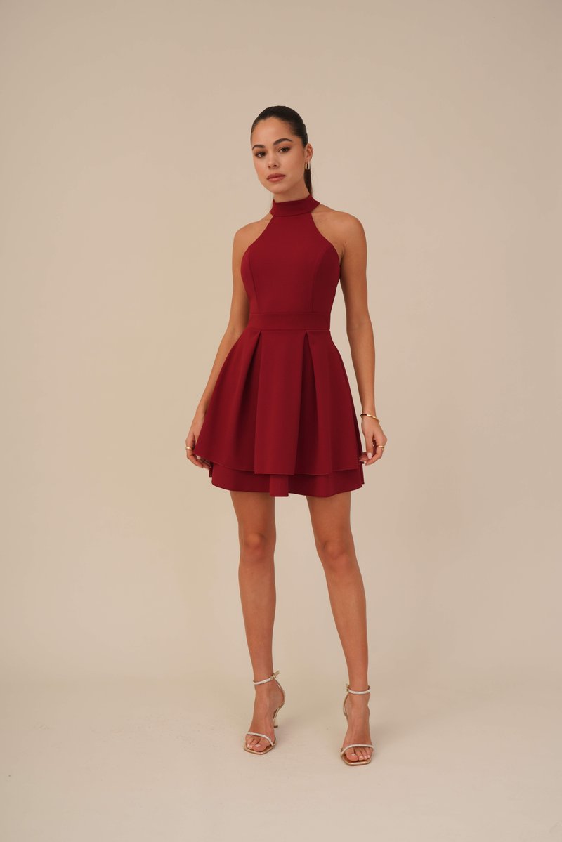 Robe rouge à col halter avec un corsage ajusté et une jupe évasée en couches. Fabriquée à partir d'un tissu texturé. Associée à des sandales à talons transparents.
