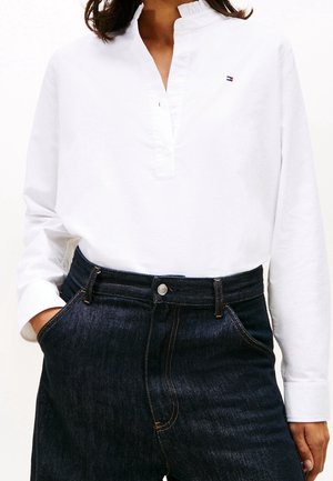 Woman wearing white long-sleeve shirt with Mandarin collar and dark high-waisted jeans, hands in pockets.