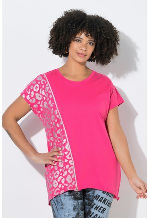 Kvinde med krøllet hår iført øreringe i ringe og en lyserød top med korte ærmer med sølv leopardprint og lynlåsdetalje, kombineret med printede jeans.