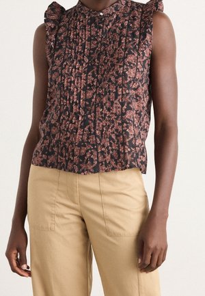 Blouse noire sans manches avec motif floral rose et épaules à volants, associée à un pantalon beige taille haute porté par une personne à la peau brun moyen.