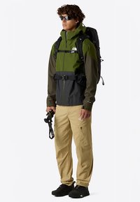 Vesitiivis, vihreä-musta takki, jossa säädettävä vyötärö ja huppu, yhdistetty vaaleanruskeisiin housuihin ja mustiin kenkiin; pitää kädessään kameraa.