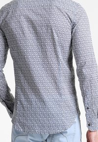 Långärmad knäppskjorta i lätt tyg med ett geometriskt mönster av blå cirklar på en vit bakgrund. Rundad nederkant och manschetter.