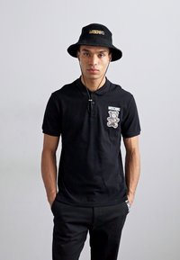 Black polo shirt with a Moschino bear logo on the left chest, paired with black pants and a matching black bucket hat.