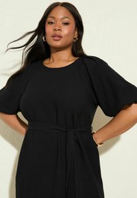 Schwarzes Kleid mit kurzen Puffärmeln, taillierter Taille und Bindegürtel-Detail. Hergestellt aus einem glatten Stoff mit minimalistischem Design und lockerer Form.
