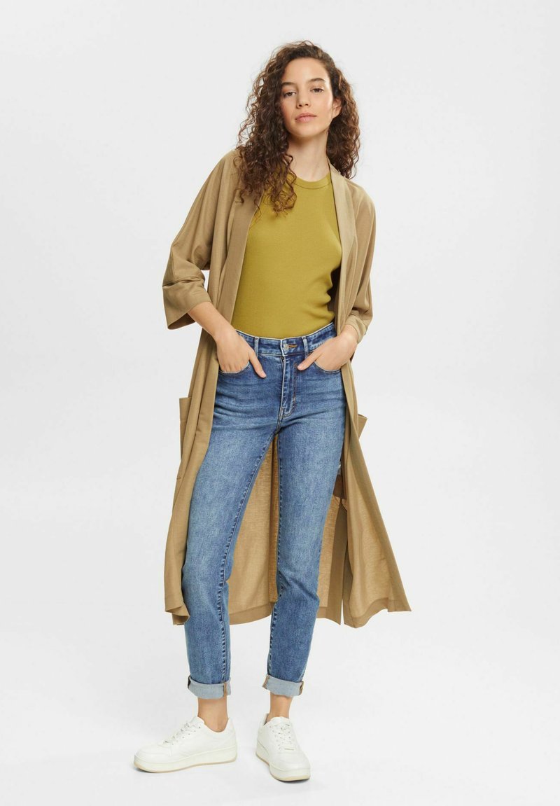 Beiger langer Cardigan über einem grünen Oberteil, kombiniert mit blauen Jeans und weißen Sneakers. Der Cardigan hat weite Ärmel und vordere Taschen.