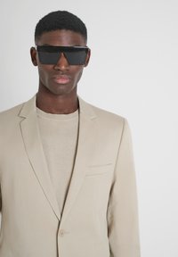 Blazer sartorial beige con rever a punta, bottone singolo e taschino sul petto, indossato sopra un maglione beige chiaro, completato da occhiali da sole neri.