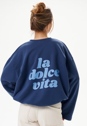 Femme avec les cheveux attachés en chignon portant un sweat-shirt bleu marine avec le texte « la dolce vita » dans le dos et un pantalon beige clair, vue de dos.