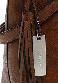 Dettaglio di una borsa in pelle marrone con un pendente, etichetta in metallo e cuciture nere a contrasto. Texture liscia con accenti hardware lucidi.