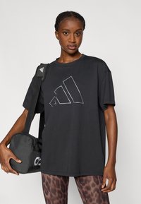 Μαύρο βαμβακερό t-shirt με λευκό γεωμετρικό λογότυπο, στρογγυλή λαιμόκοψη και κοντά μανίκια, σε συνδυασμό με μαύρη τσάντα και κολάν με σχέδια.