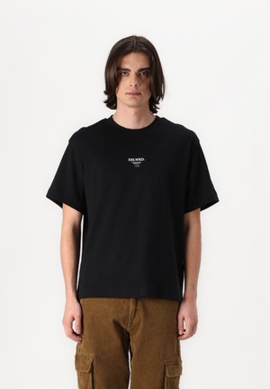 Schwarzes Baumwoll-T-Shirt mit kurzen Ärmeln und Rundhalsausschnitt. Zentrales weißes Textlogo auf der Brust. Getragen mit braunen Cordhosen.