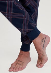 Marine joggerbroeken met een rood en blauw ruitpatroon, voorzien van elastische boorden. Blootgestelde voeten met zichtbare tatoeages op de huid.