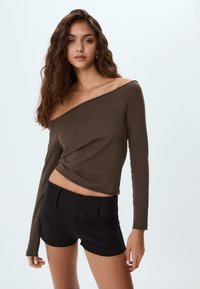 Brun off-shoulder långärmad topp med vridet framtill, kombinerad med svarta högmidjade shorts, visad mot en ljus bakgrund.