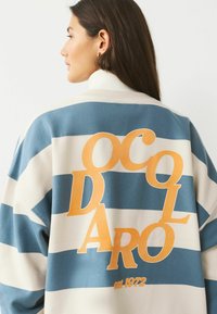 Striped sweatshirt in blue and cream with large orange graphic text on the back, featuring ribbed collar and relaxed fit.