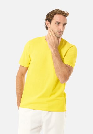 Mies keltaisessa lyhythihaisessa paidassa ja valkoisissa housuissa koskettaa leukaansa ja katsoo sivulle yksinkertaisen vaalean taustan edessä.