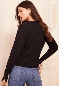 Pulover negru cu tiv ondulat și accente de butoni pe mâneci, purtat cu blugi din denim albastru. Textură moale și design ajustat.