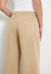 Los pantalones vaqueros anchos beige cuentan con una alta cintura, bolsillos traseros y un pequeño logo triangular rojo. La tela tiene una textura suave.