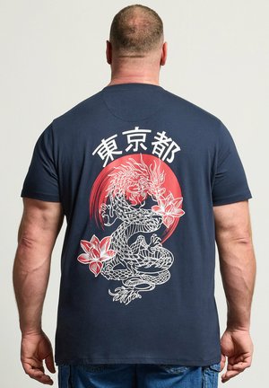 Man wearing navy blue T-shirt with white and red dragon and flower design and Japanese characters on the back, standing against plain background.