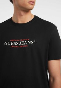 Schwarzes Baumwoll-T-Shirt mit kurzen Ärmeln und Rundhalsausschnitt. Es hat weißen und roten Text mit der Aufschrift "GUESS JEANS AMERICAN TRADITION ORIGINAL DESIGNS."