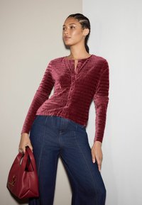 Dunkelrotes strukturiertes Langarmshirt mit Knopfverschluss und rundem Halsausschnitt, kombiniert mit blauen weit geschnittenen Jeans und einer roten Handtasche.