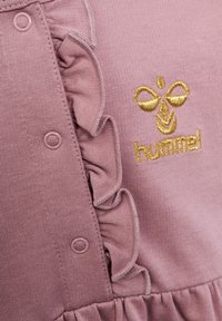 Tessuto in maglia rosa con bordino a balze, bottoni a pressione e logo a ricamo dorato di un'ape sopra al testo "hummel".