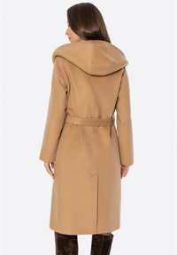 Kamelowy wełniany płaszcz z dużym kapturem, wiązaniem w talii, długimi rękawami i prostą sylwetką, z rozcięciem z tyłu dla swobody ruchu.