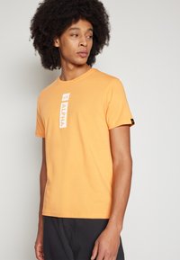 Orange bomulds t-shirt med korte ærmer, der har et vertikalt hvidt "ALPHA" logo. Stoffet har en glat tekstur og en normal pasform.