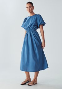 Ženska v modri midi obleki s pouf puff rokavi in elastičnim pasom, v kombinaciji s črnimi ravnimi sandali na trakove, ki stoji nasproti enobarvne podlage.