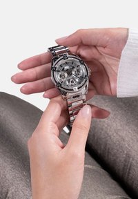 Srebrna kronograf ura s tremi poddiali, kristalni okraski na okvirju, kovinski pas in prozorno zunanje steklo.