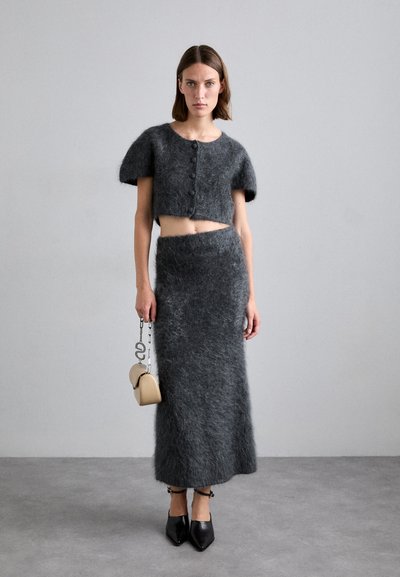 Donna che indossa un cardigan corto grigio scuro in pelliccia e una gonna lunga abbinata, tiene una borsa beige con tracolla a catena e scarpe nere con punta.